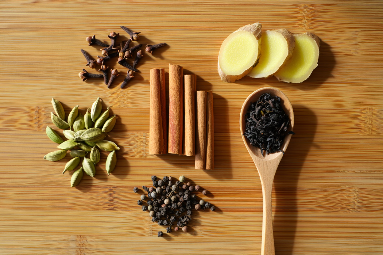 Whole chai spices and loose Assam CTC black tea on light oak wood: green cardamom pods, cinnamon sticks, fresh ginger, cloves, and black peppercorns